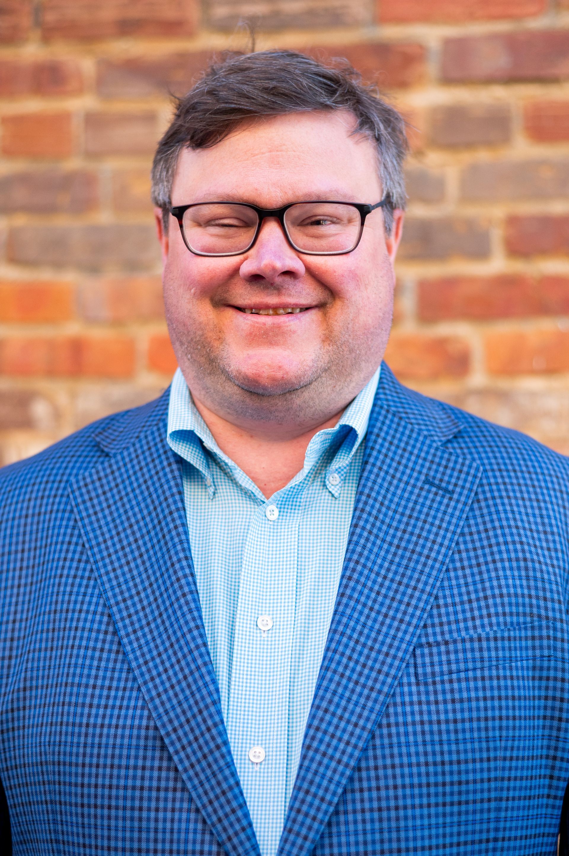 Man with glasses smiles, wearing blue blazer and light blue shirt, brick wall background.
