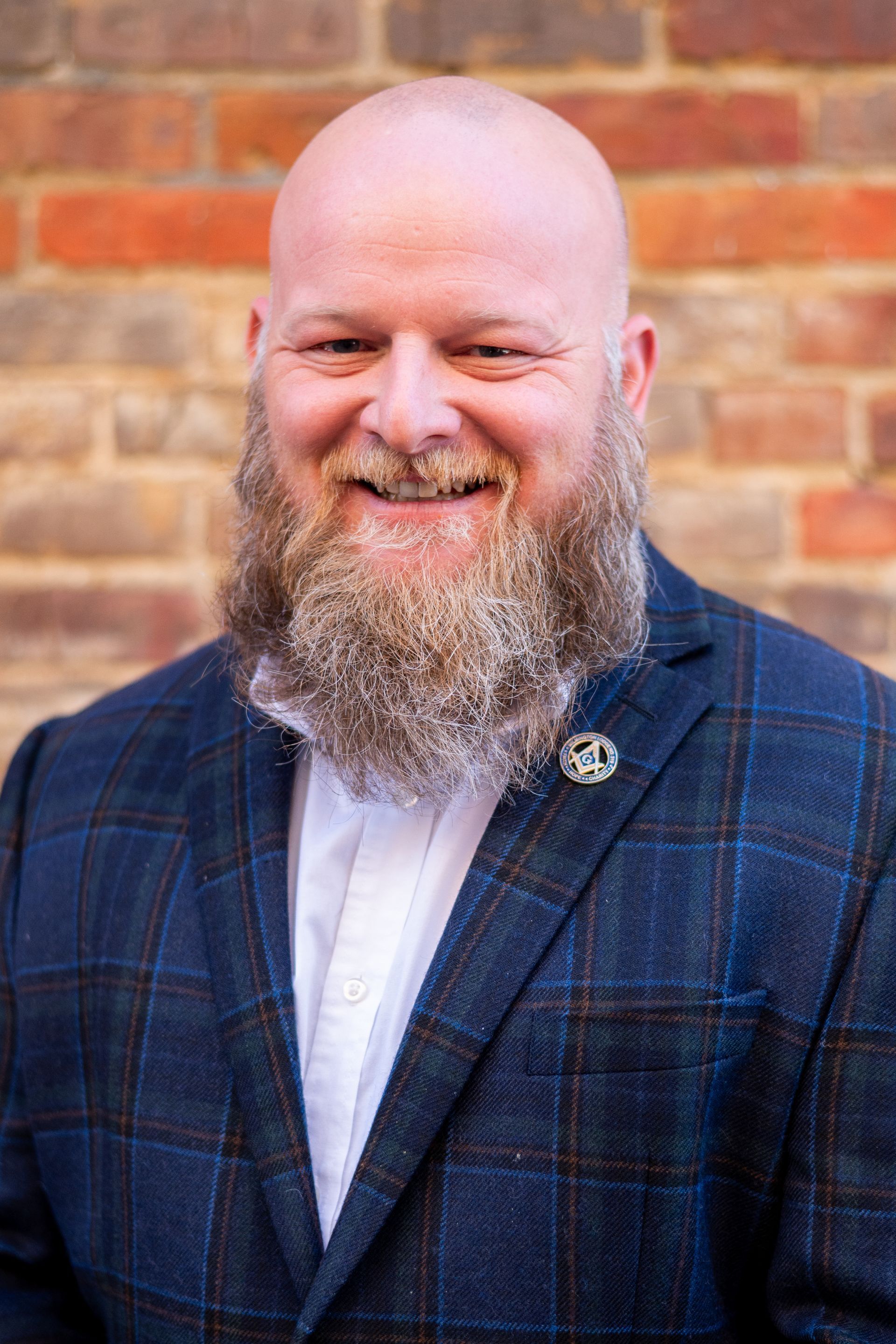 Man with bald head and long beard in plaid blazer smiles in front of a brick wall.