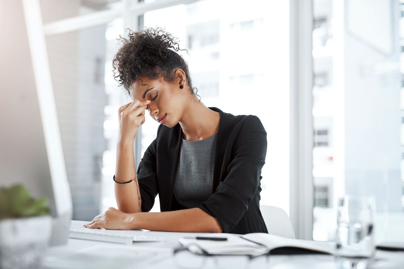 Person seated at a desk, rubbing their forehead while looking down at papers in a bright office