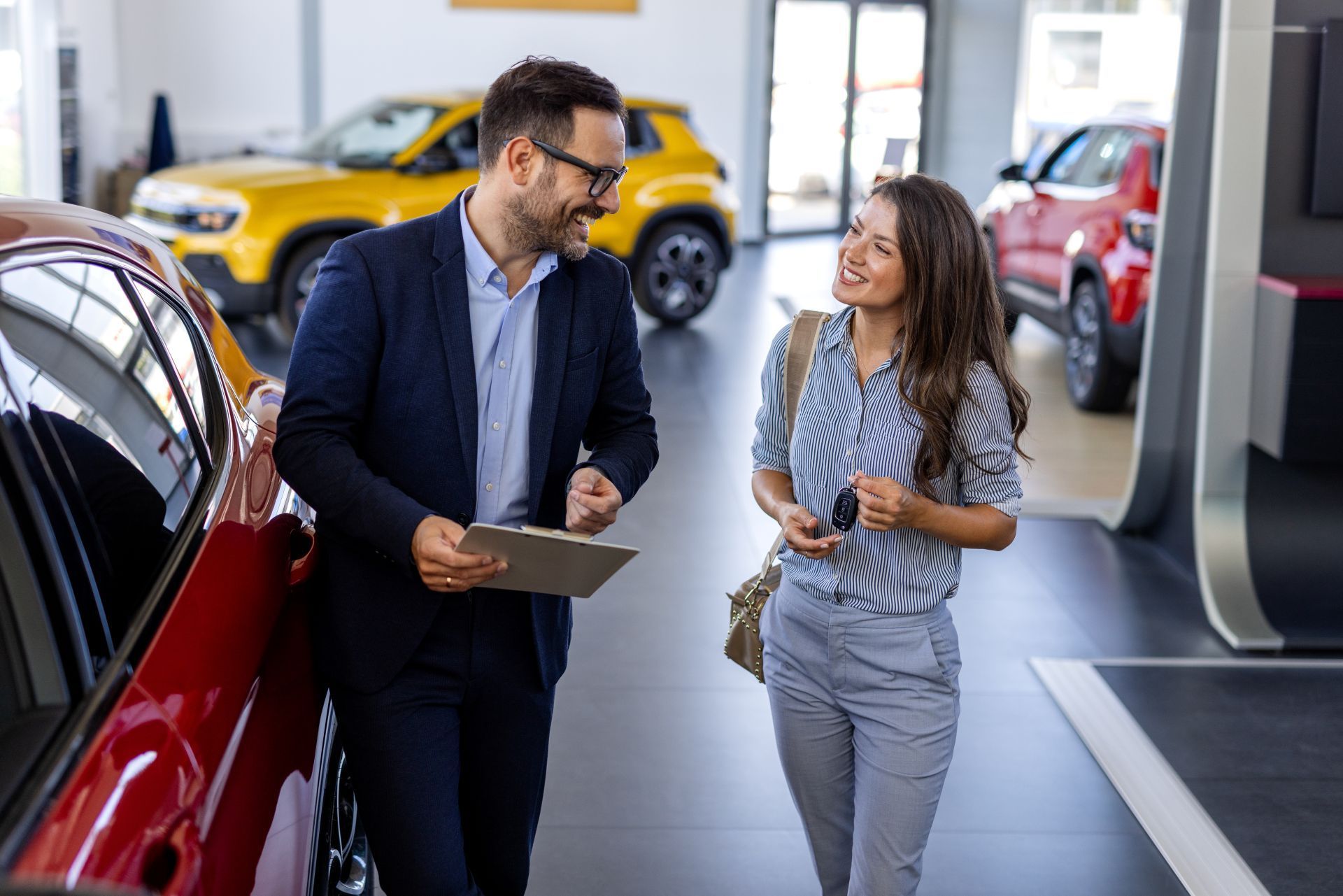 Two people talking in a car dealership showroom beside a red car, with yellow and red cars in the background