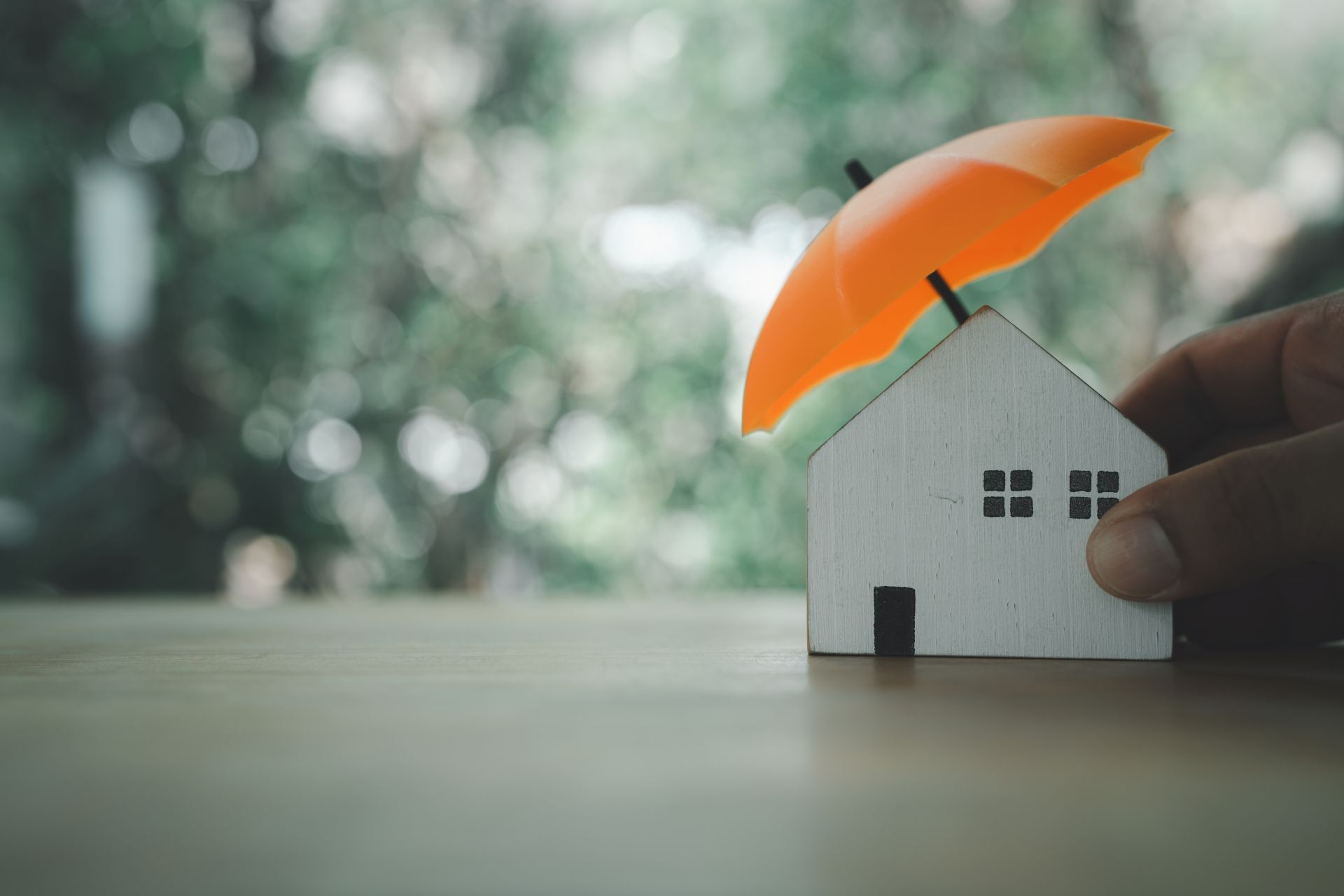 Hand holding a small house model with an orange umbrella on top, symbolizing home protection