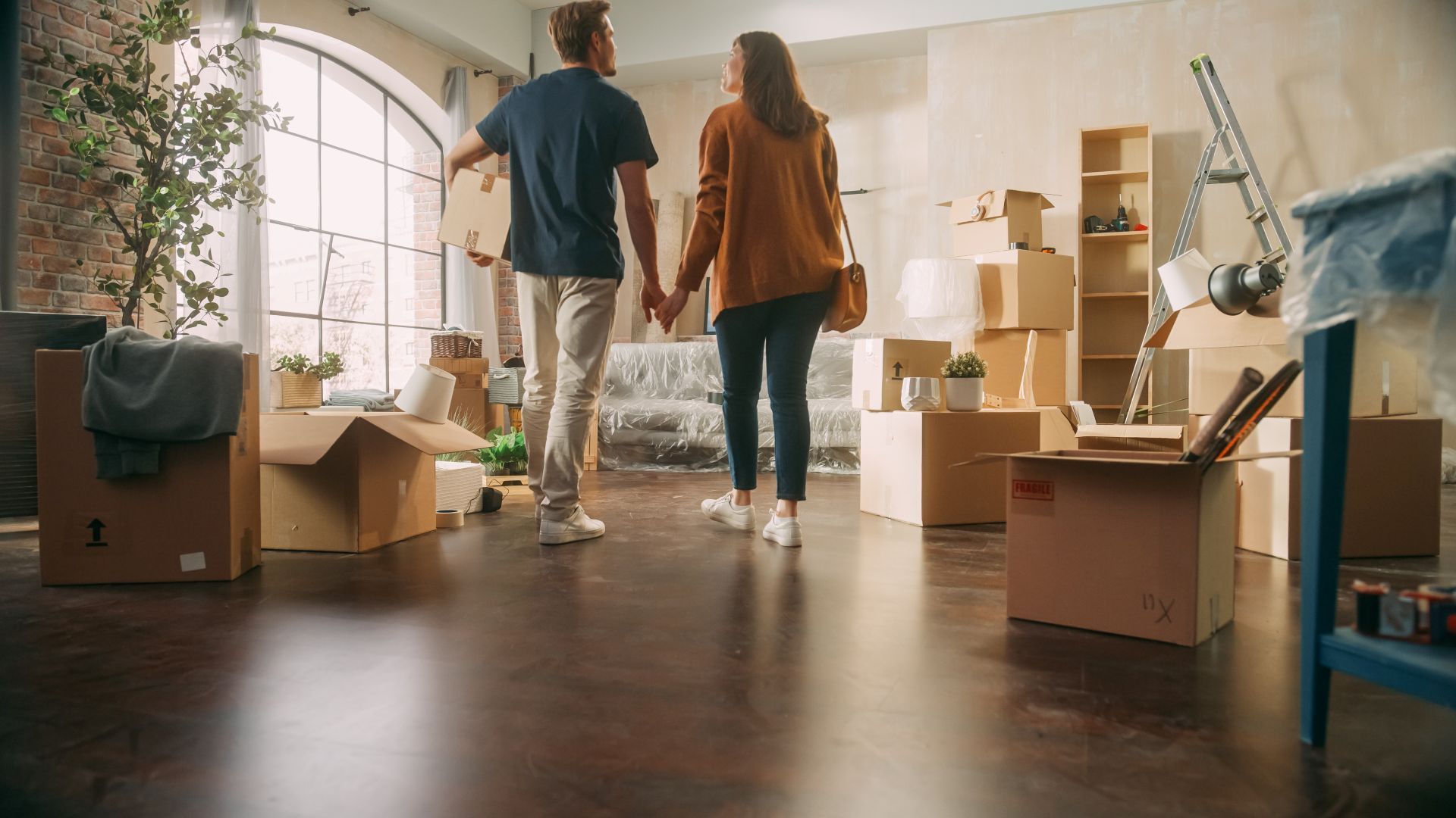 Two people walk through a sunlit room filled with moving boxes and packed furniture.