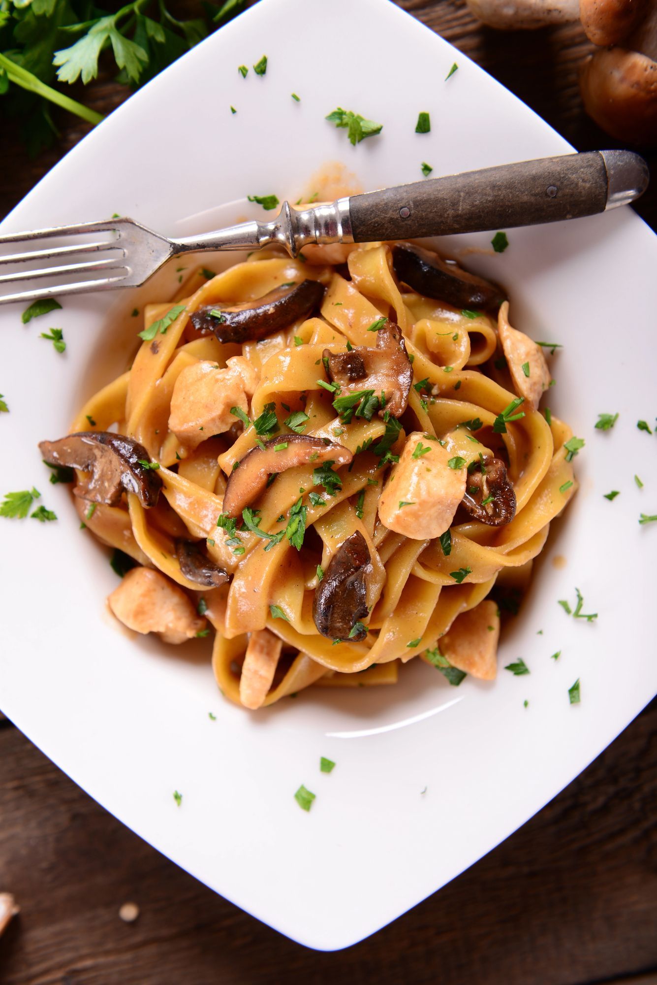 Piatto di pasta con funghi, pollo e prezzemolo in una ciotola quadrata bianca con una forchetta.