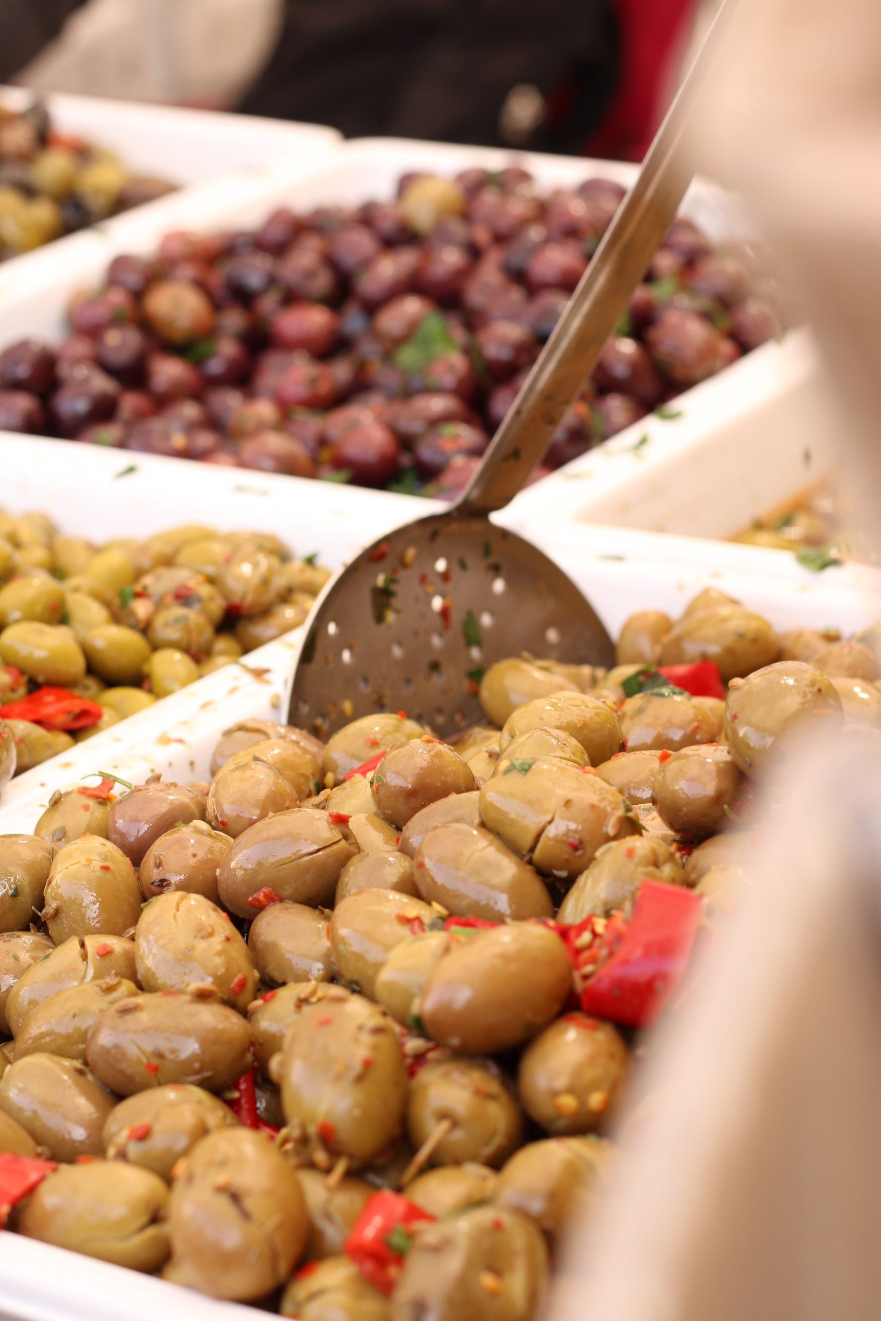 Olive di vari colori, tra cui verde e viola, al mercato. Un mestolo forato estrae le olive da un contenitore.