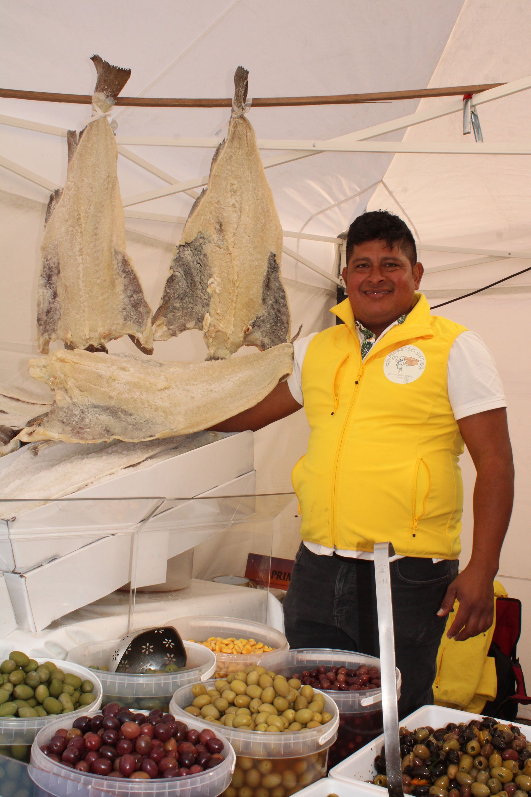 Un uomo con il gilet giallo è in piedi davanti a una bancarella del mercato con olive e pesce essiccato.