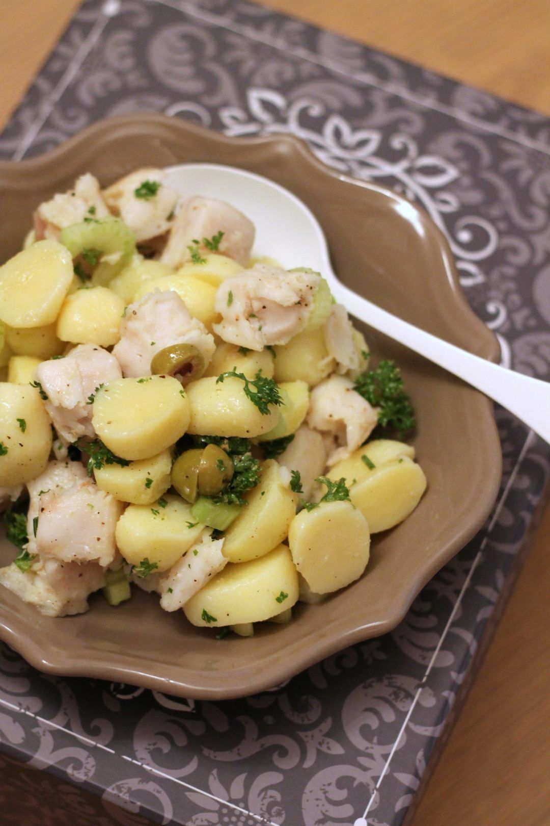 Insalata di patate con pesce, olive ed erbe aromatiche in una ciotola marrone con un cucchiaio bianco su una tovaglietta a fiori.