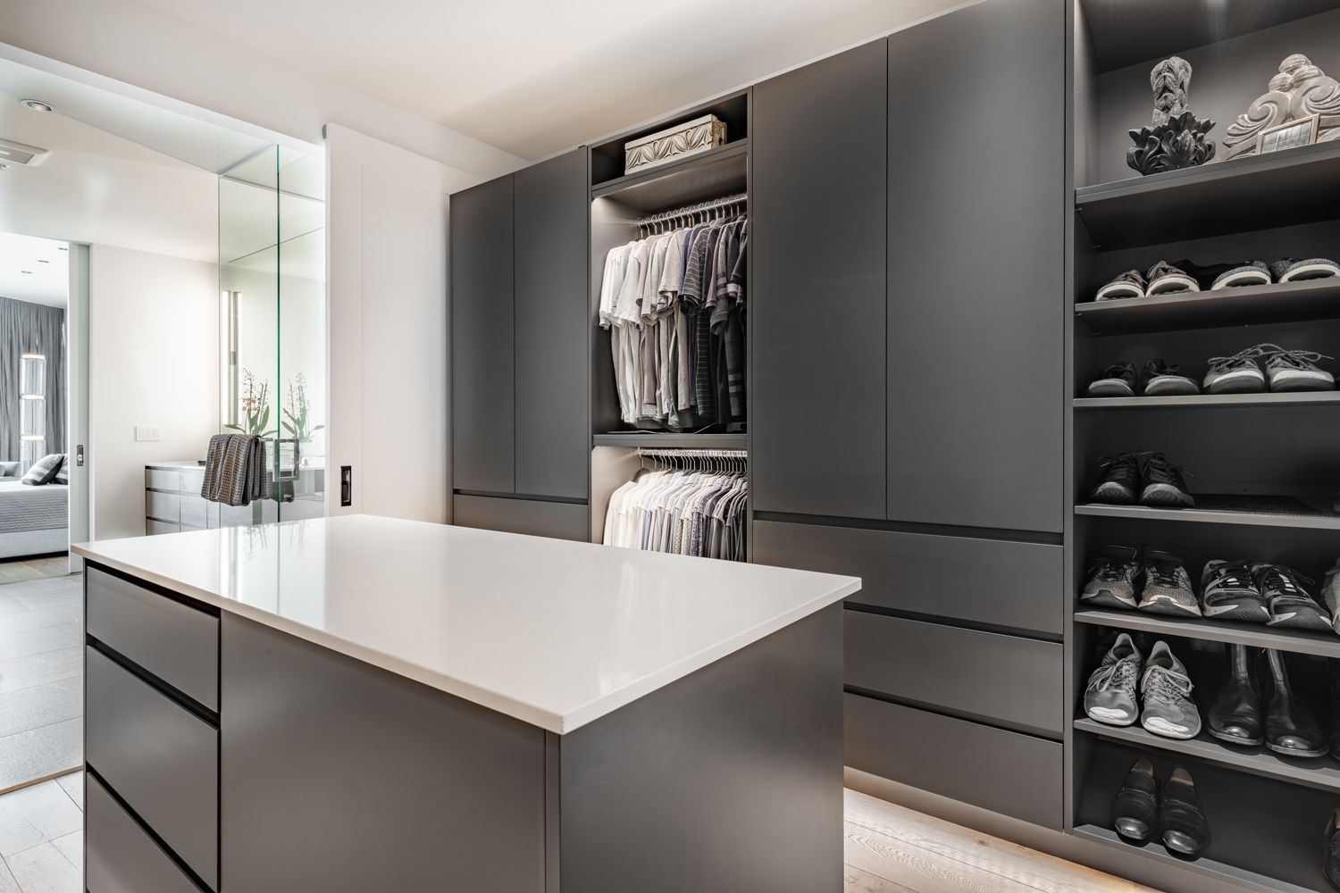 Modern gray walk-in closet with island, open shelving for shoes, and sliding glass door to bedroom.