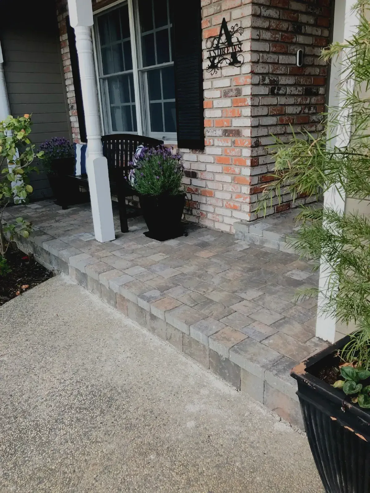 Brick patio leading to a house entrance with a black bench, potted plants, and a white column.