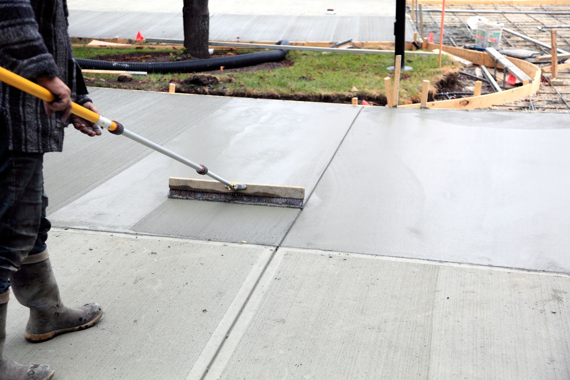 Person smoothing wet concrete with a long-handled tool on a sidewalk. Construction site with grass and forms in the background.