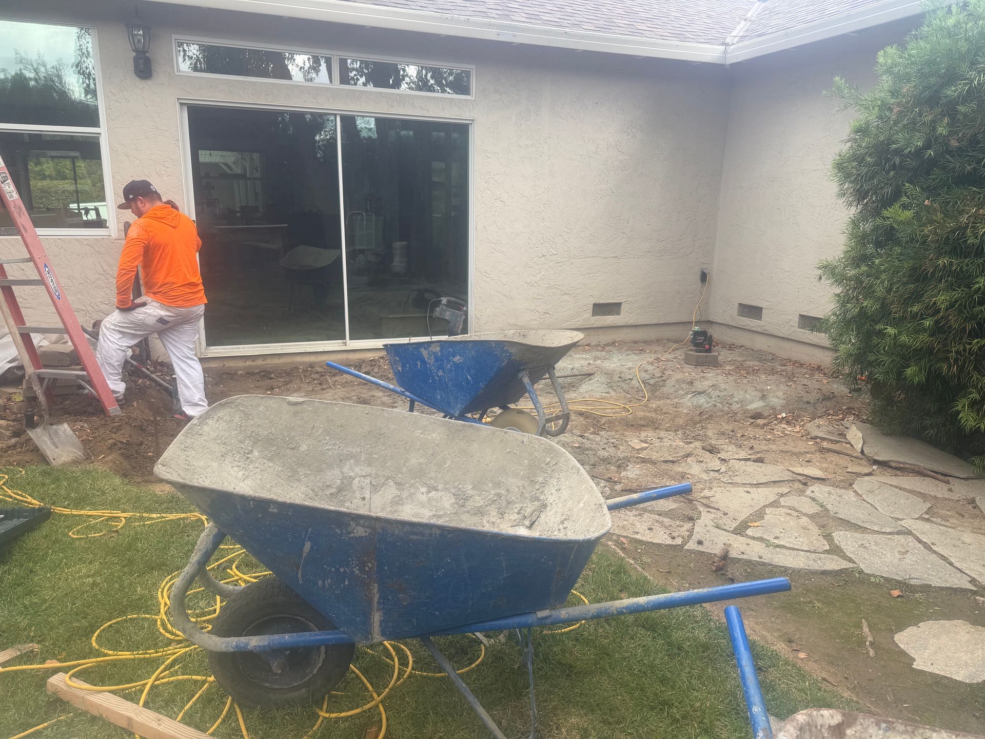 Construction site. A man in an orange jacket works near a sliding glass door. Two blue wheelbarrows are visible.