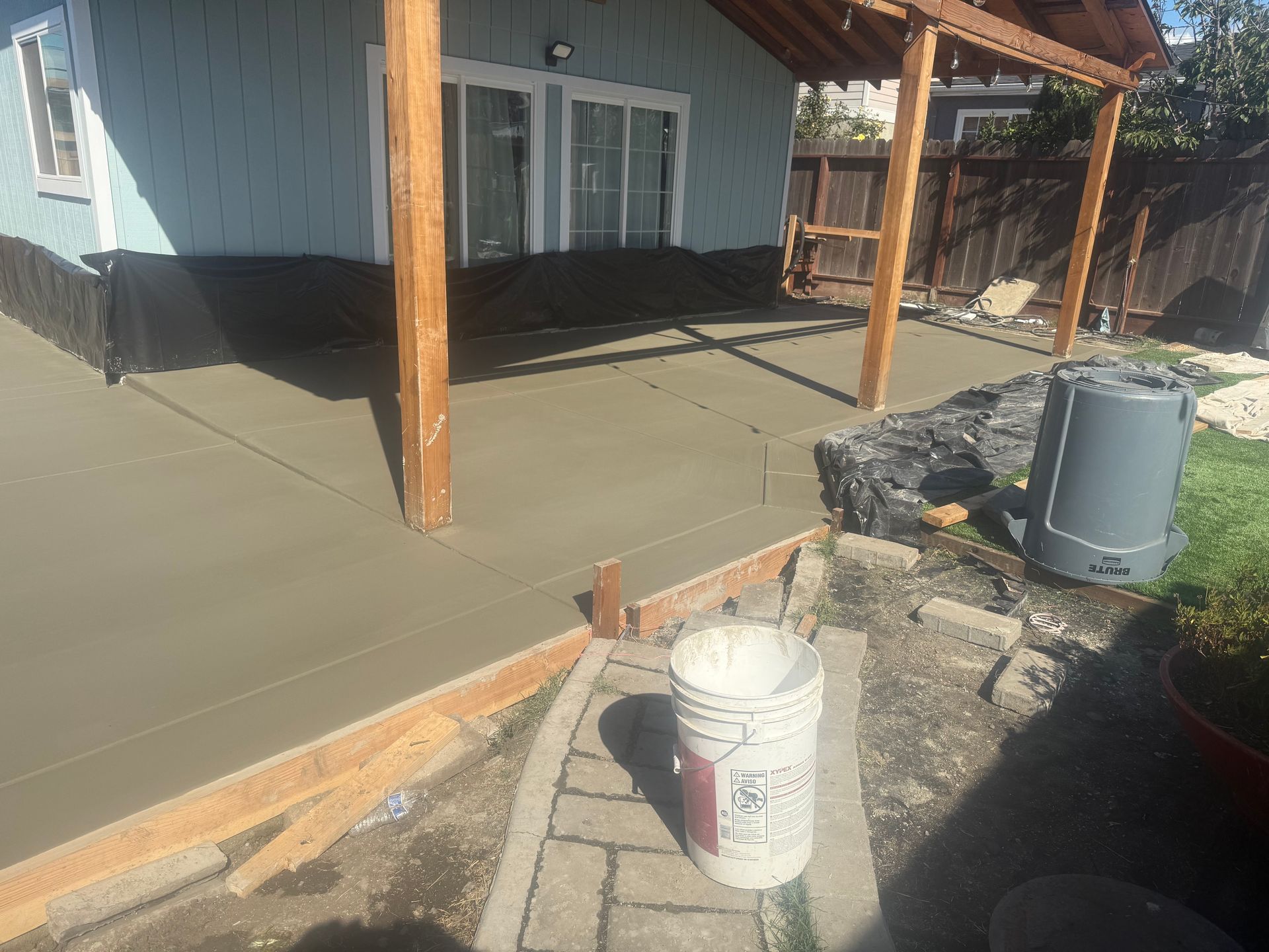 Newly poured concrete patio next to a light blue house with a wooden pergola. A bucket sits in foreground.