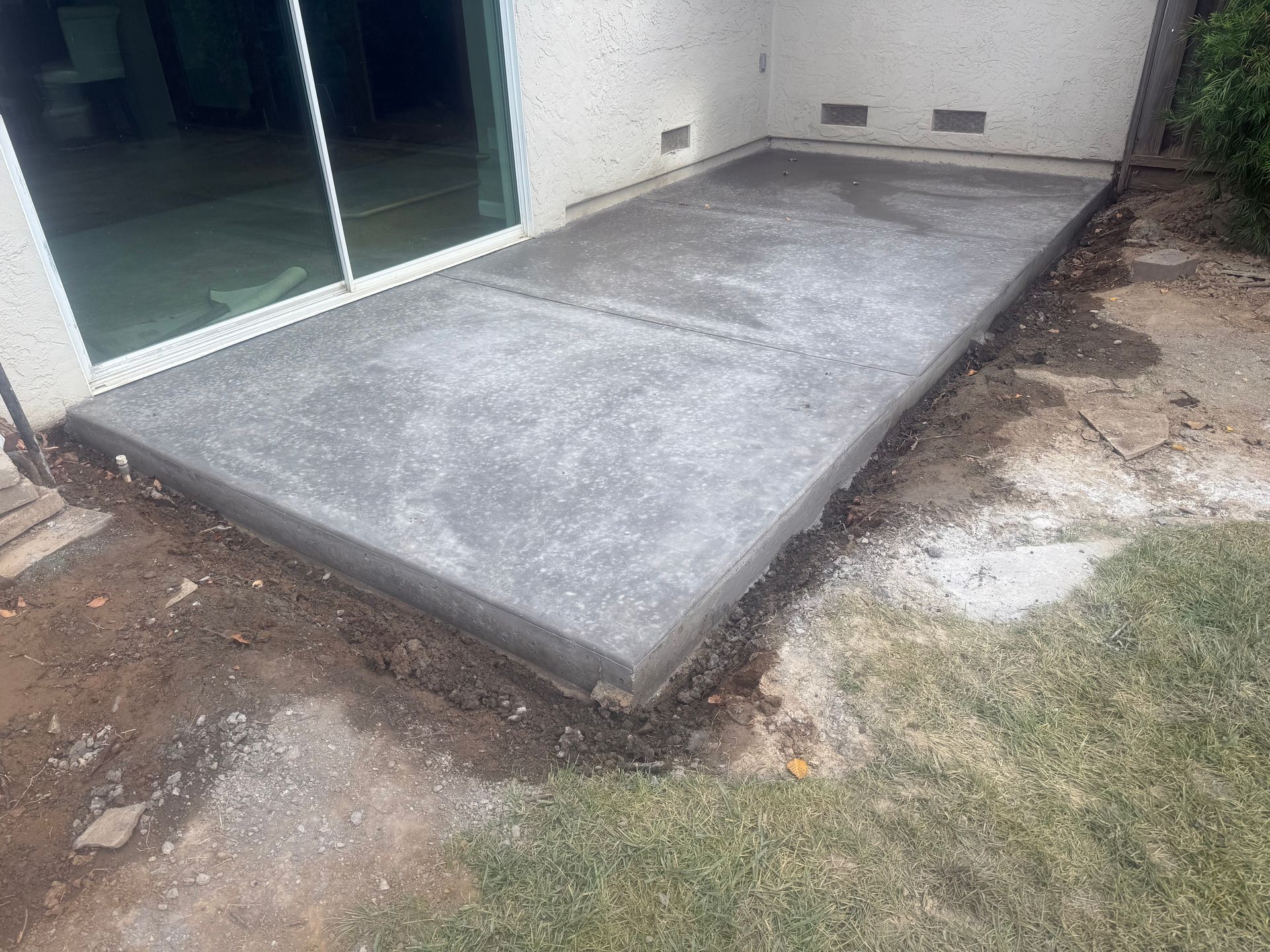 Concrete patio next to a sliding glass door. The ground is dirt and grass.