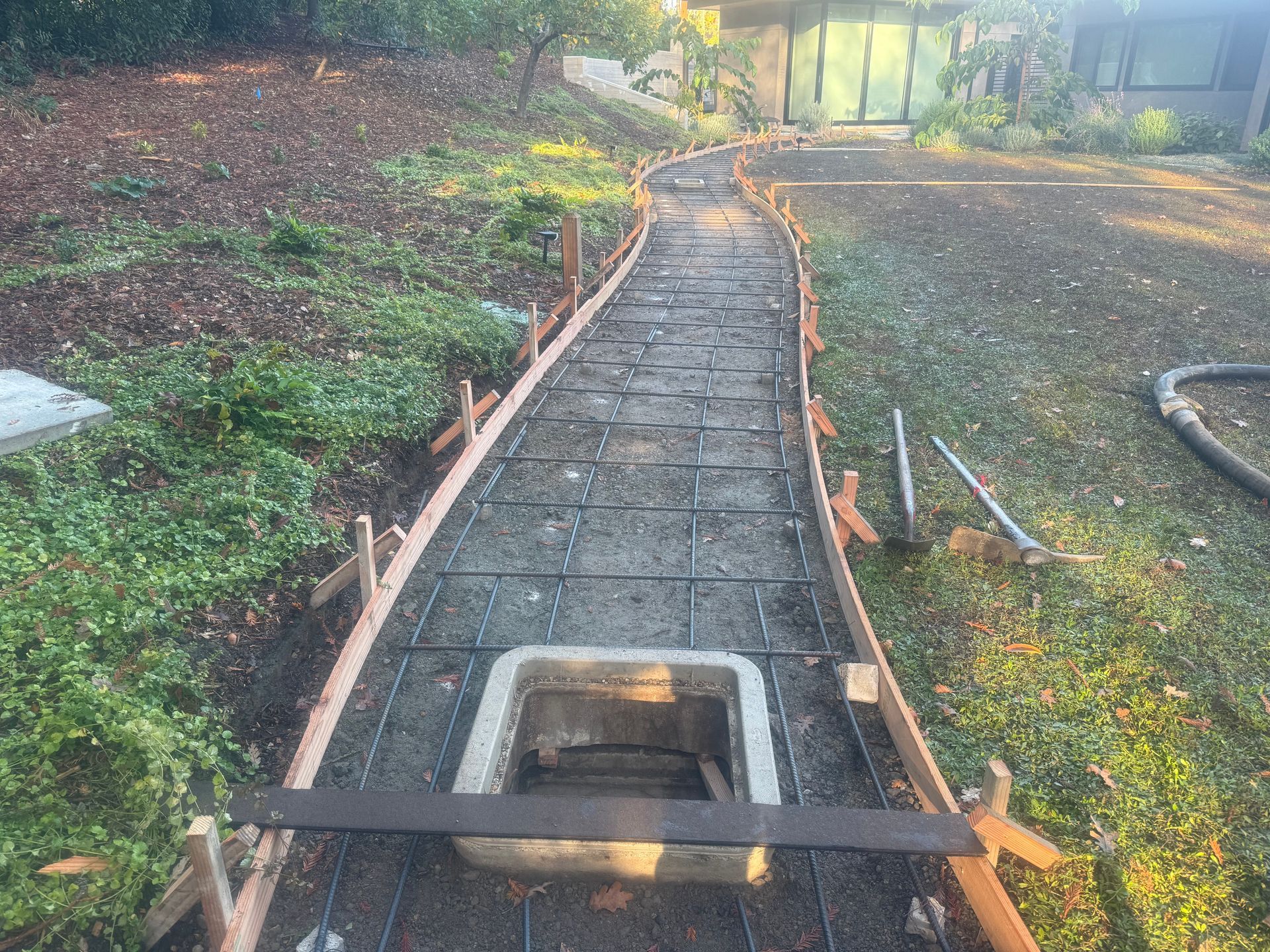 Pathway construction: wooden forms, rebar grid, a square utility access, green grass, and hillside.
