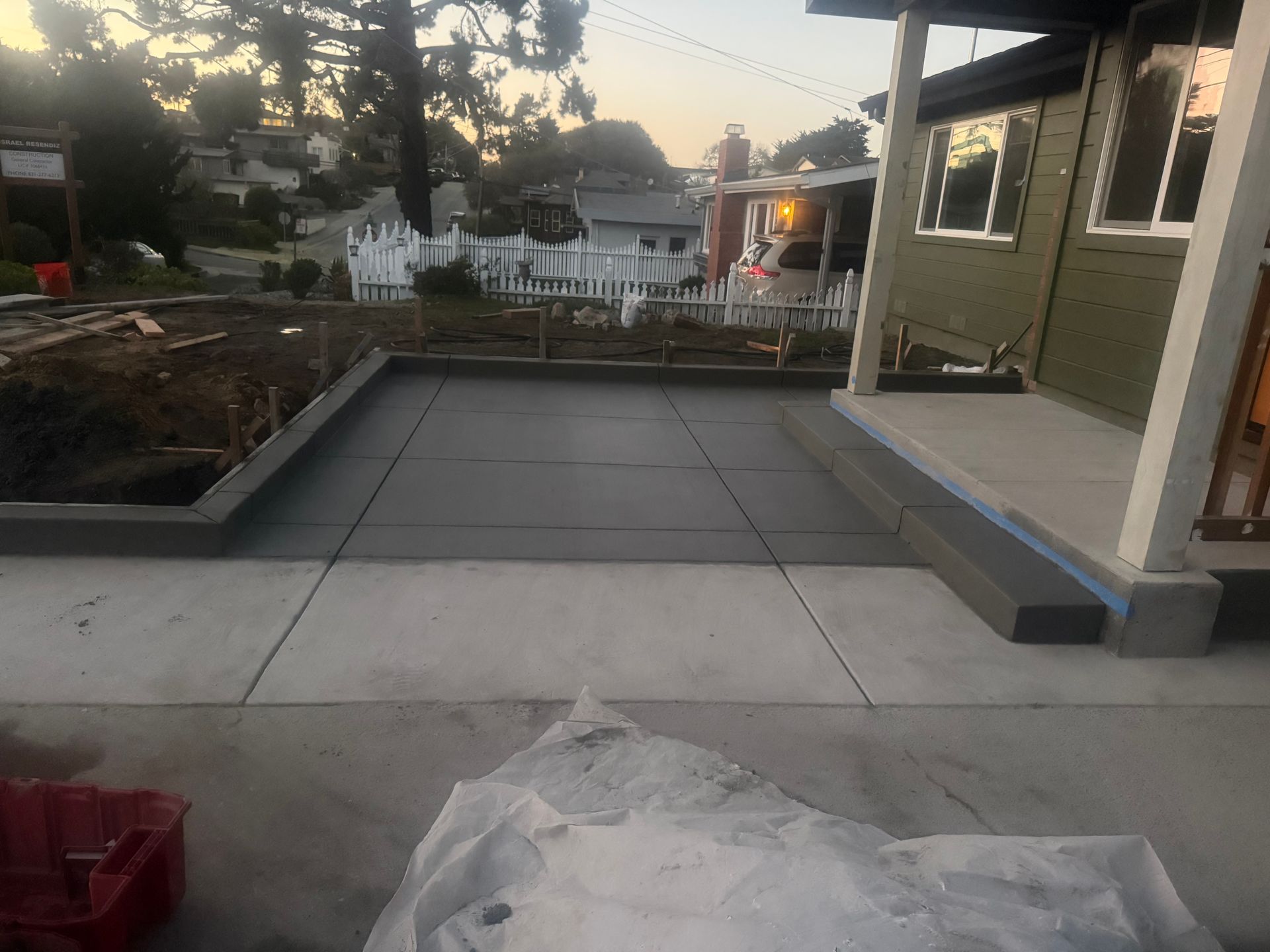 Newly poured concrete patio and walkway in front of a house, with a garden bed on the left and a white picket fence in the background.