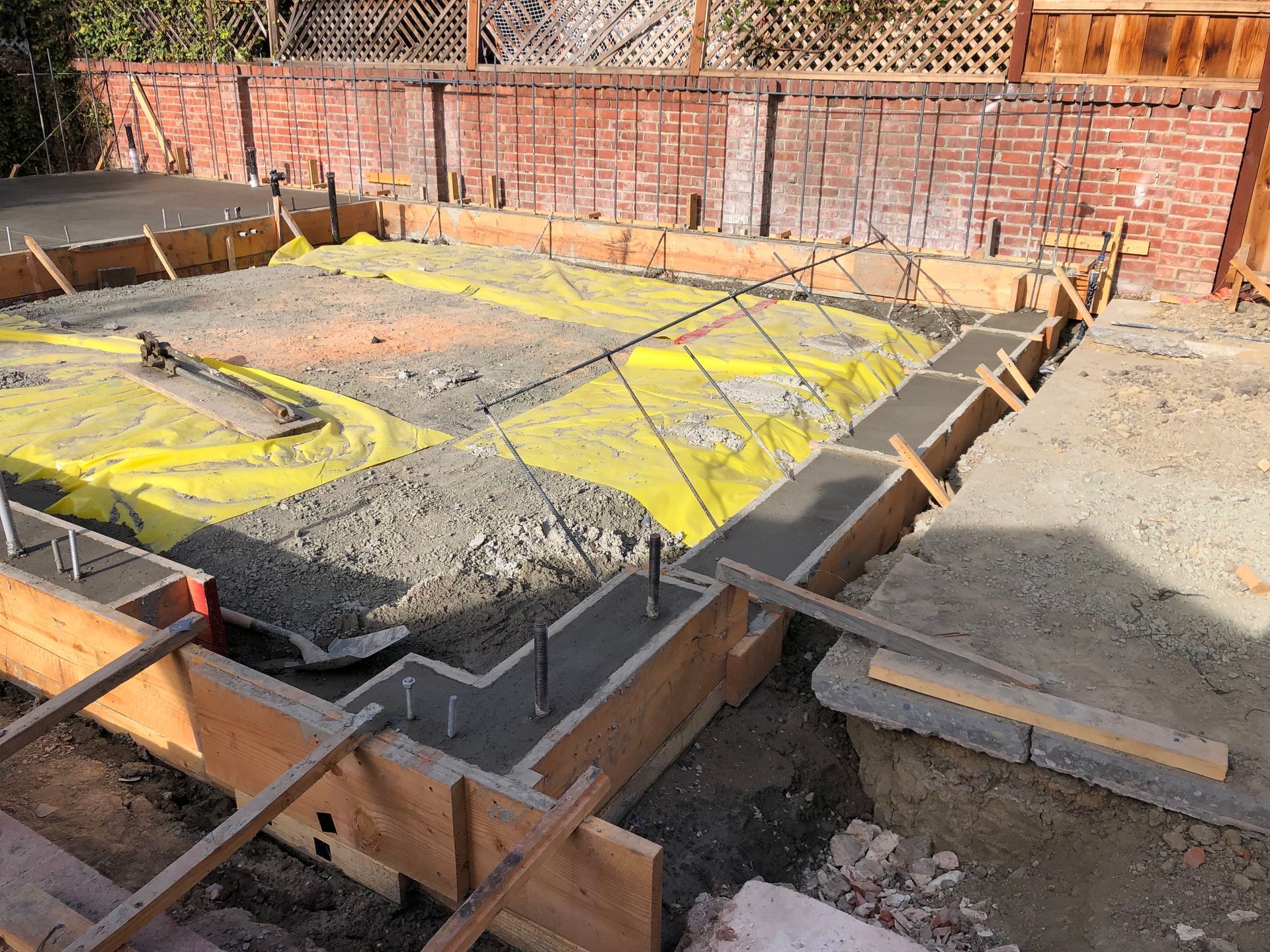 Construction site: wooden forms outline a concrete foundation, rebar, and gravel. Yellow sheeting covers the ground.