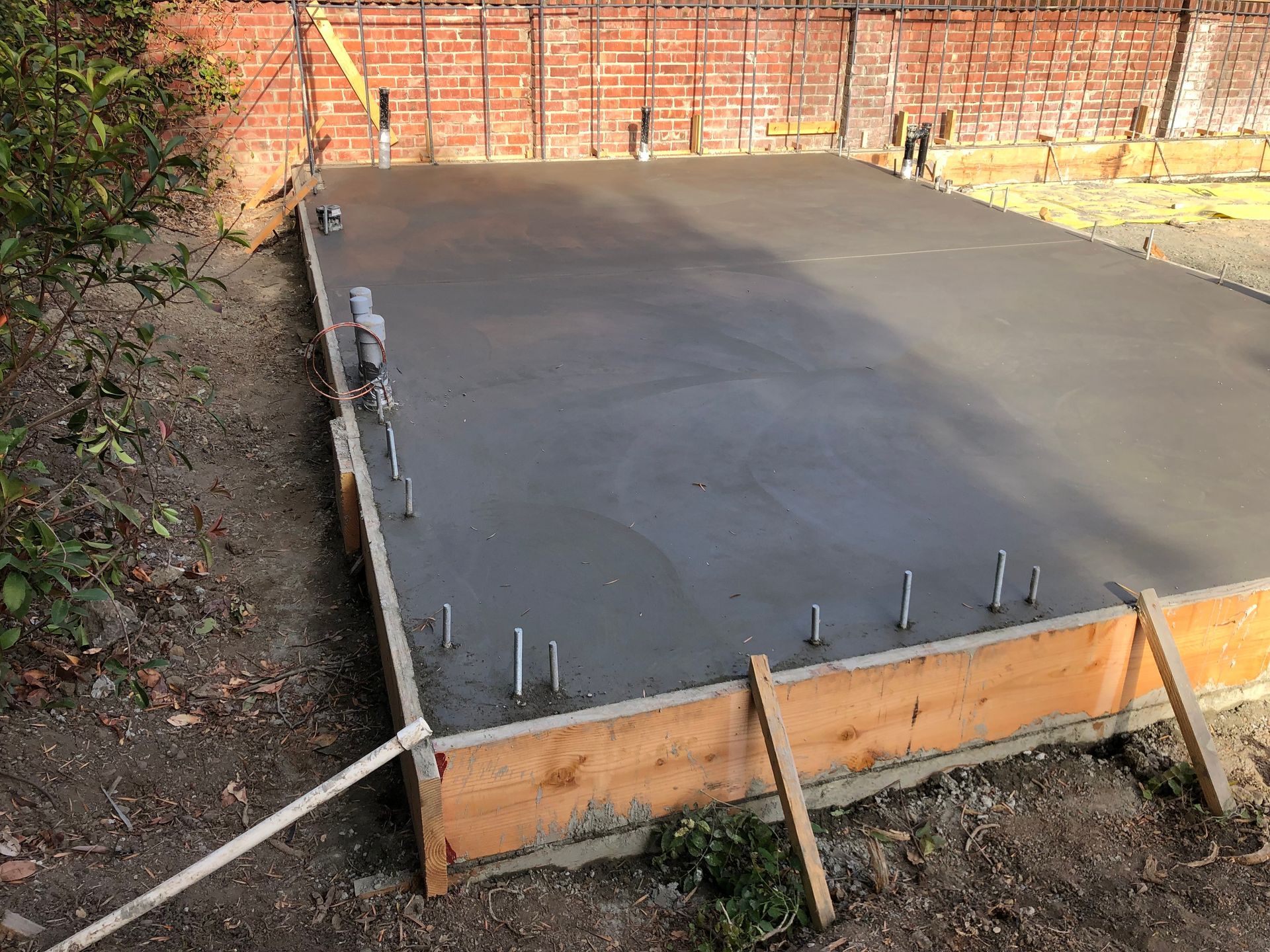 Freshly poured concrete slab within wooden formwork, set in a backyard. Steel rods protrude from the surface near brick wall.