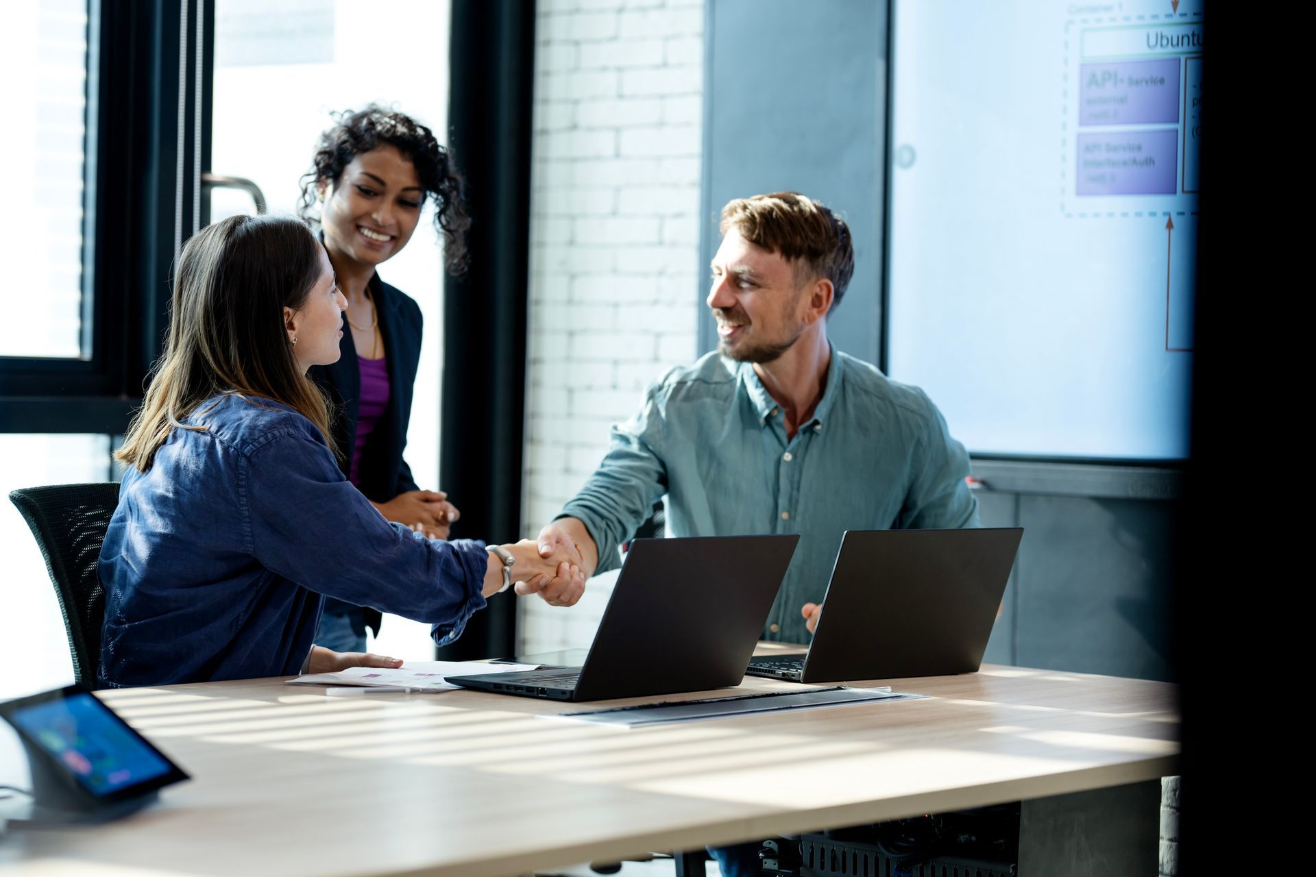 Three people in an office shake hands over laptops, a woman smiles behind them.