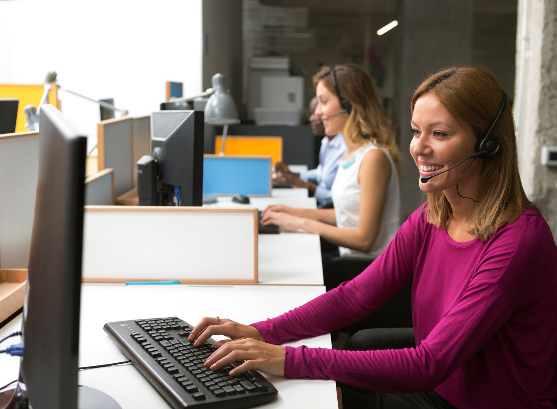 Woman with headset smiling, typing at computer in an office, other colleagues in background.