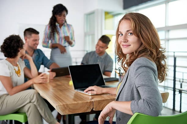 Woman smiles at camera while working on laptop at a table with a group of colleagues in an office setting.
