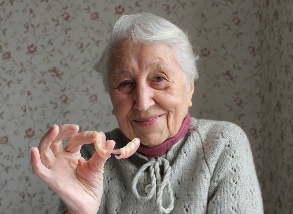 Elderly person holding a set of dentures, smiling at the camera. Light-colored sweater against a floral wallpaper backdrop.
