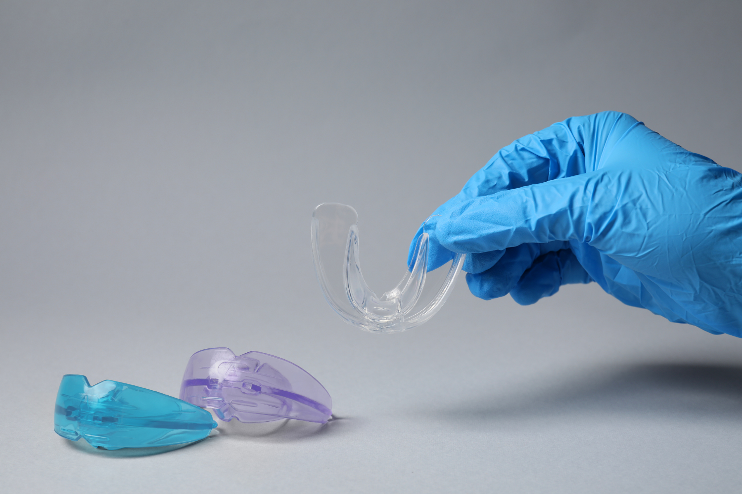 Gloved hand holding a clear dental retainer above two colored molds on a gray surface.
