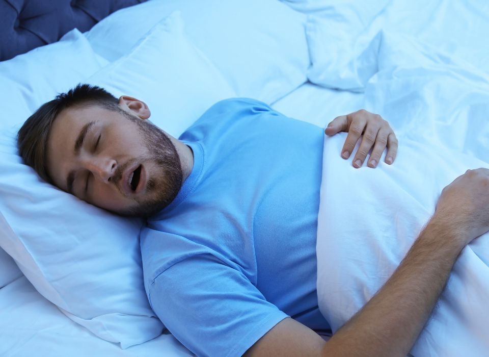 Man sleeping in a bed, mouth open, covered in white sheets, blue shirt.
