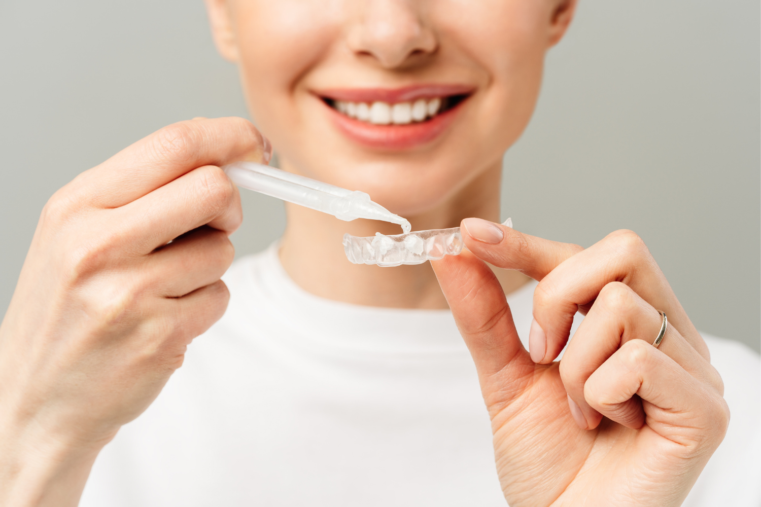 Woman smiles, applying product to clear aligner. Gray background, white shirt.