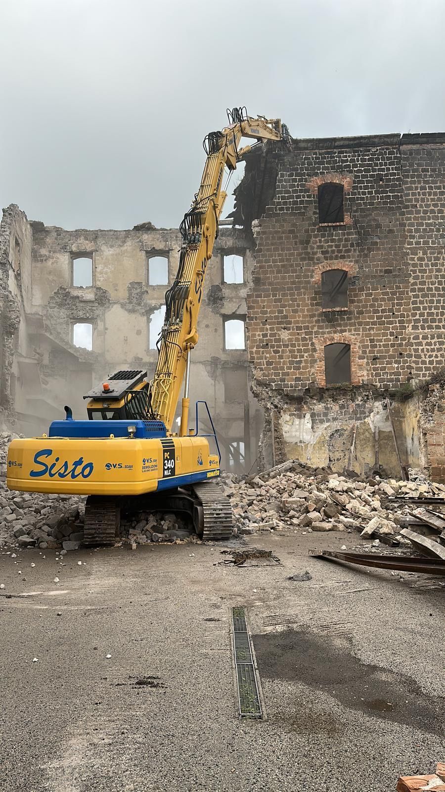 Un escavatore giallo e blu sta demolendo un edificio in mattoni.
