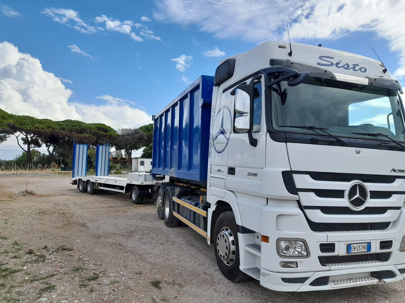 Un camion Mercedes bianco con un rimorchio blu è parcheggiato in un campo sterrato.