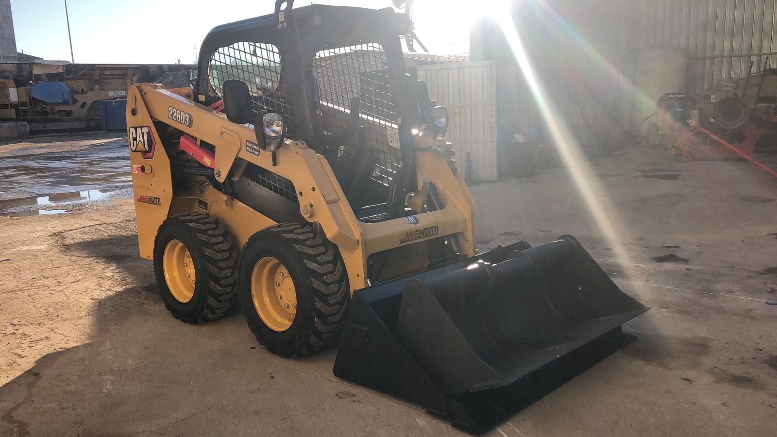 Una pala compatta gialla e nera con una grande benna è parcheggiata in un piazzale sterrato.