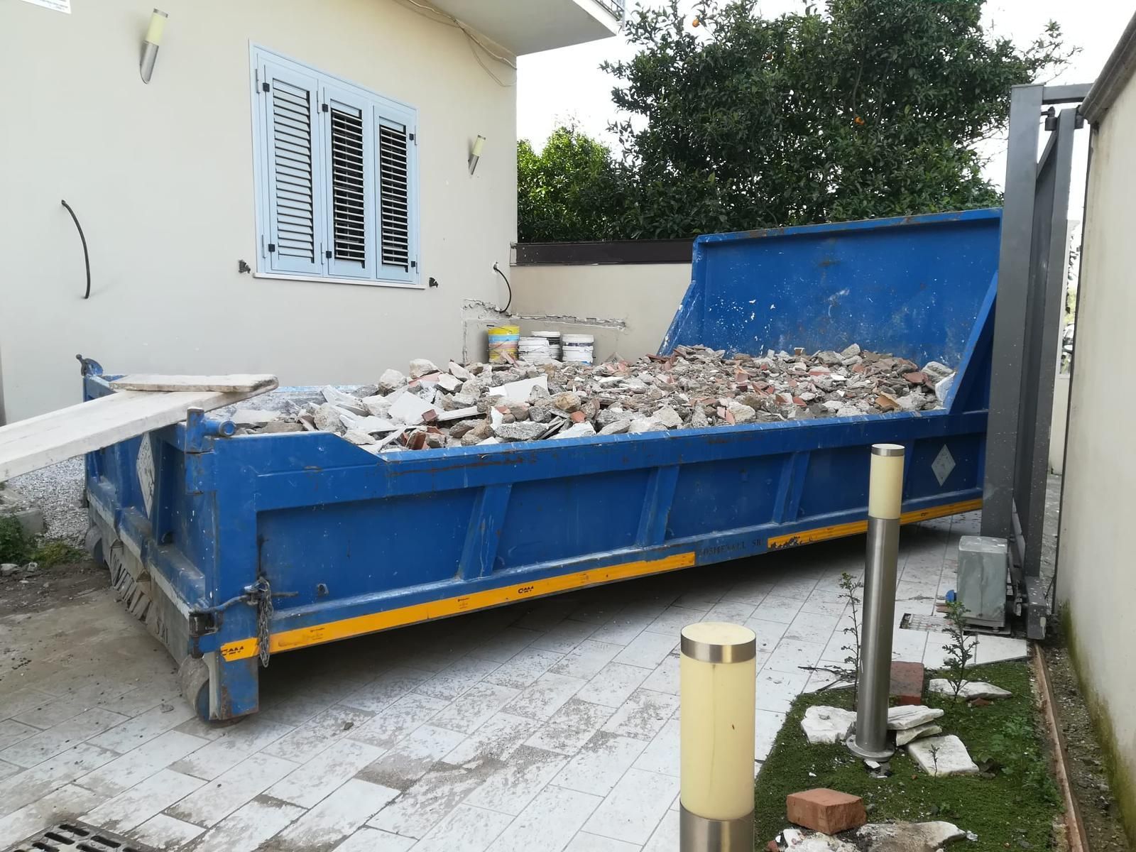 Un cassonetto blu pieno di mattoni è parcheggiato davanti a una casa.