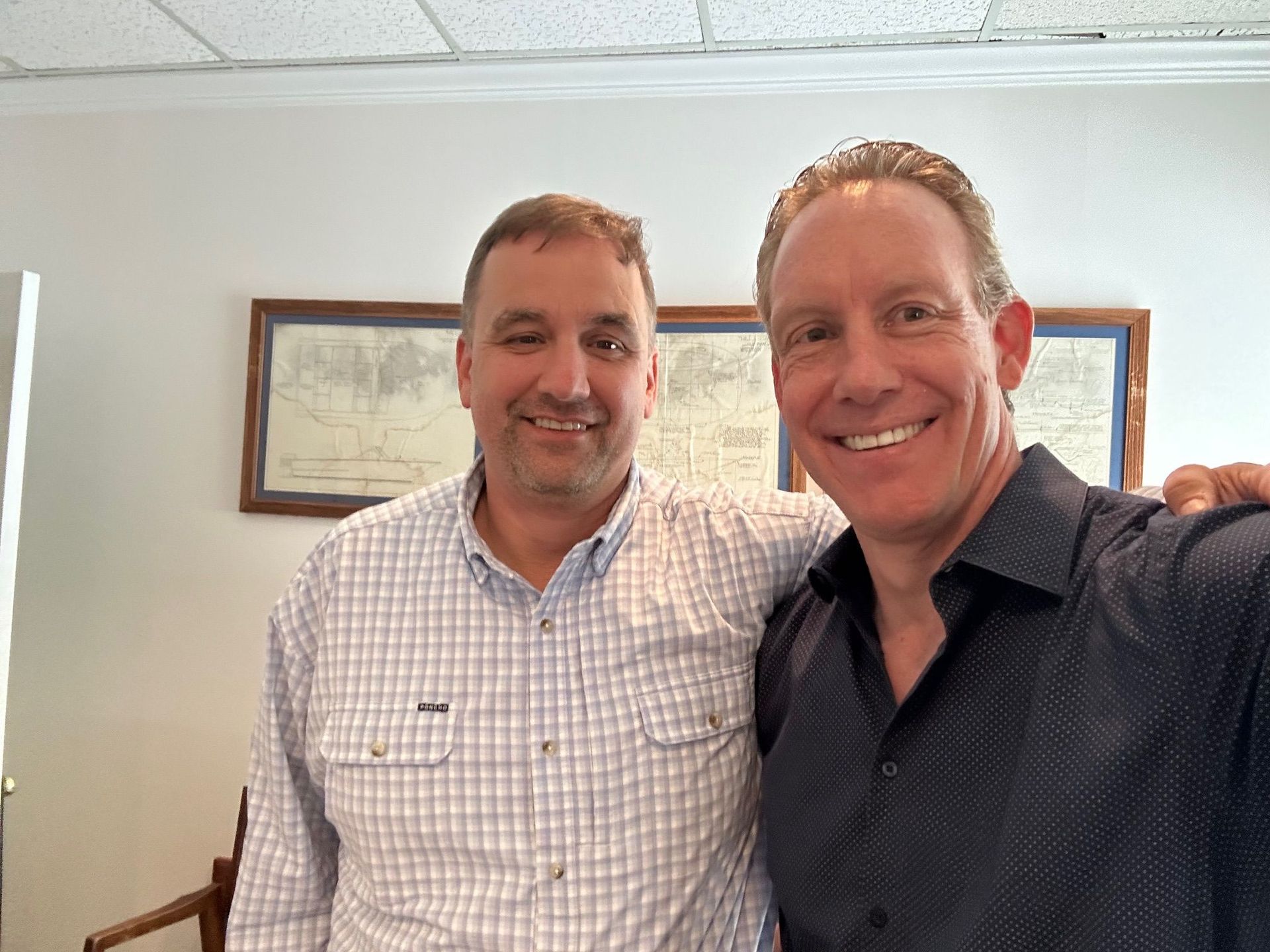 Two men smile, arm-in-arm, inside a room with framed artwork. One wears a checkered shirt; the other, a dark button-down.