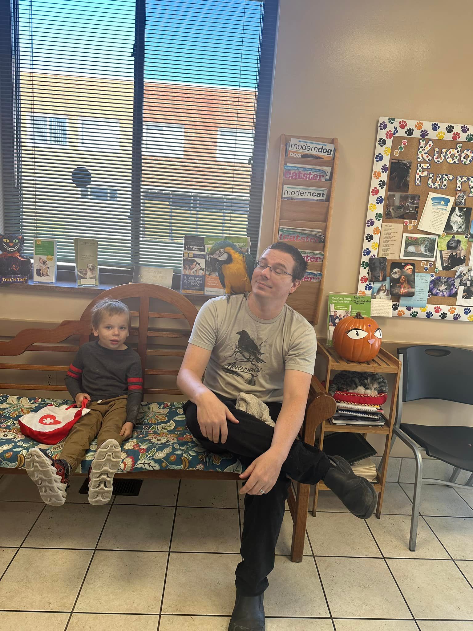 Man with a macaw on his shoulder sits on a bench next to a child, indoors near a window.