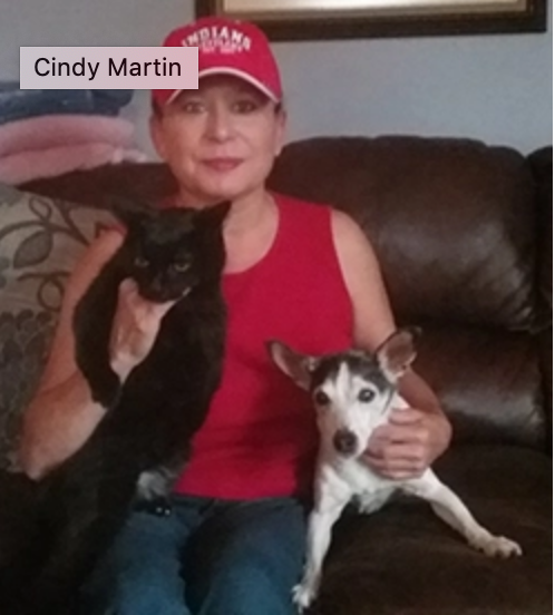 Woman in red top and hat sits on a couch, holding a black cat and a small dog.