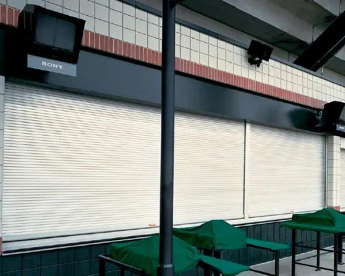 Closed storefront with white roller shutters, green table covers, and a 