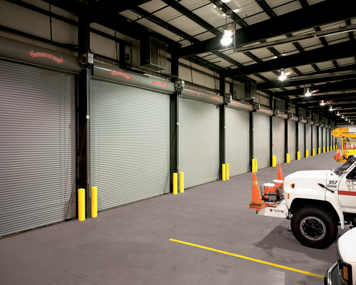 Industrial warehouse with metal roll-up doors, yellow bollards, and parked utility vehicles.