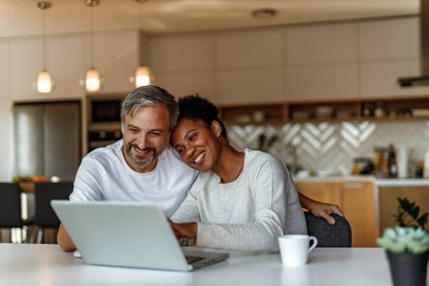 Smiling couple, white man and Black woman, look at a laptop in a modern kitchen.