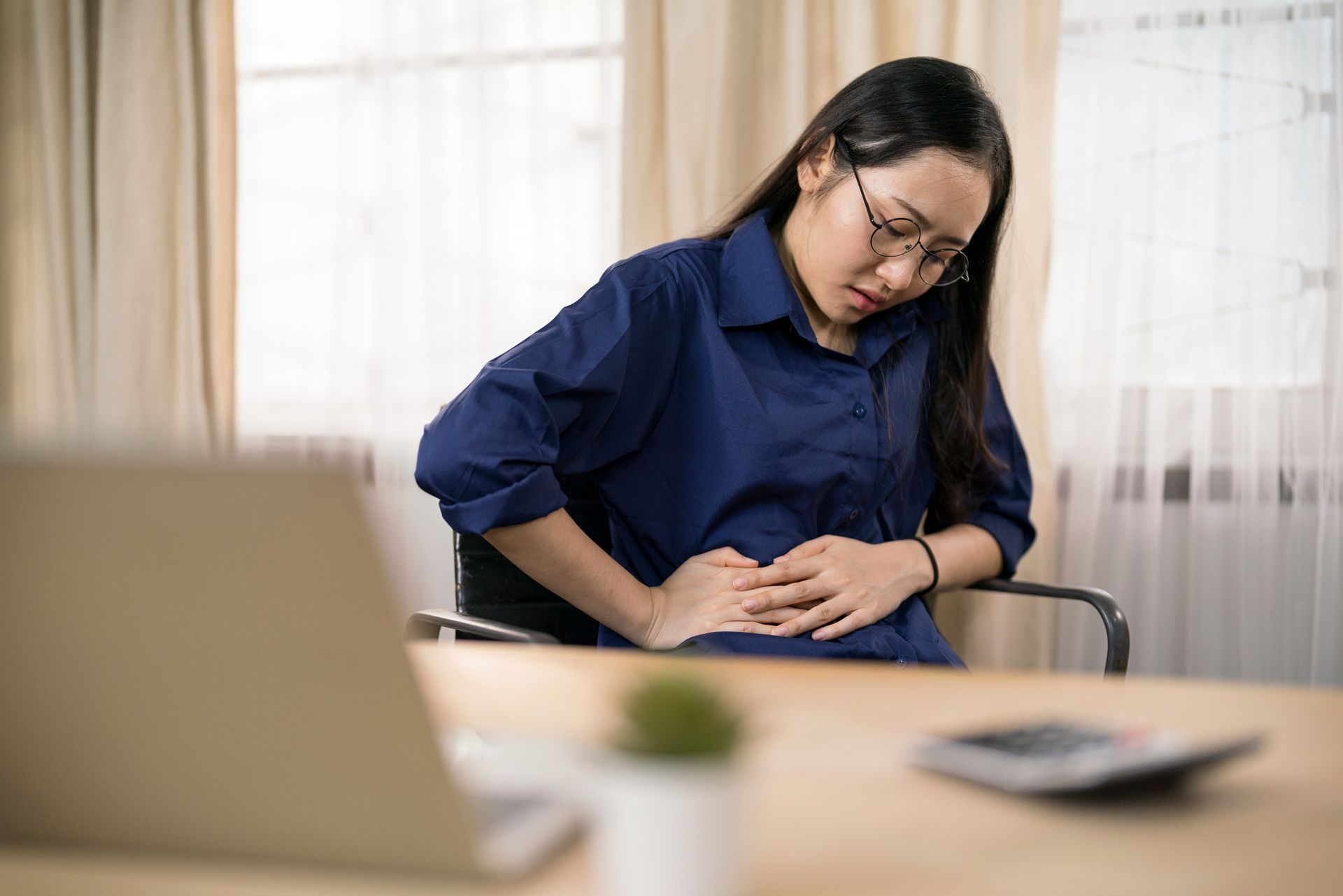 Woman with glasses, in blue shirt, holds stomach, seated at desk with laptop, appearing in pain.