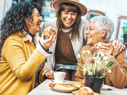 Three laughing women at an outdoor cafe, one holding a croissant, enjoying coffee and each other's company.