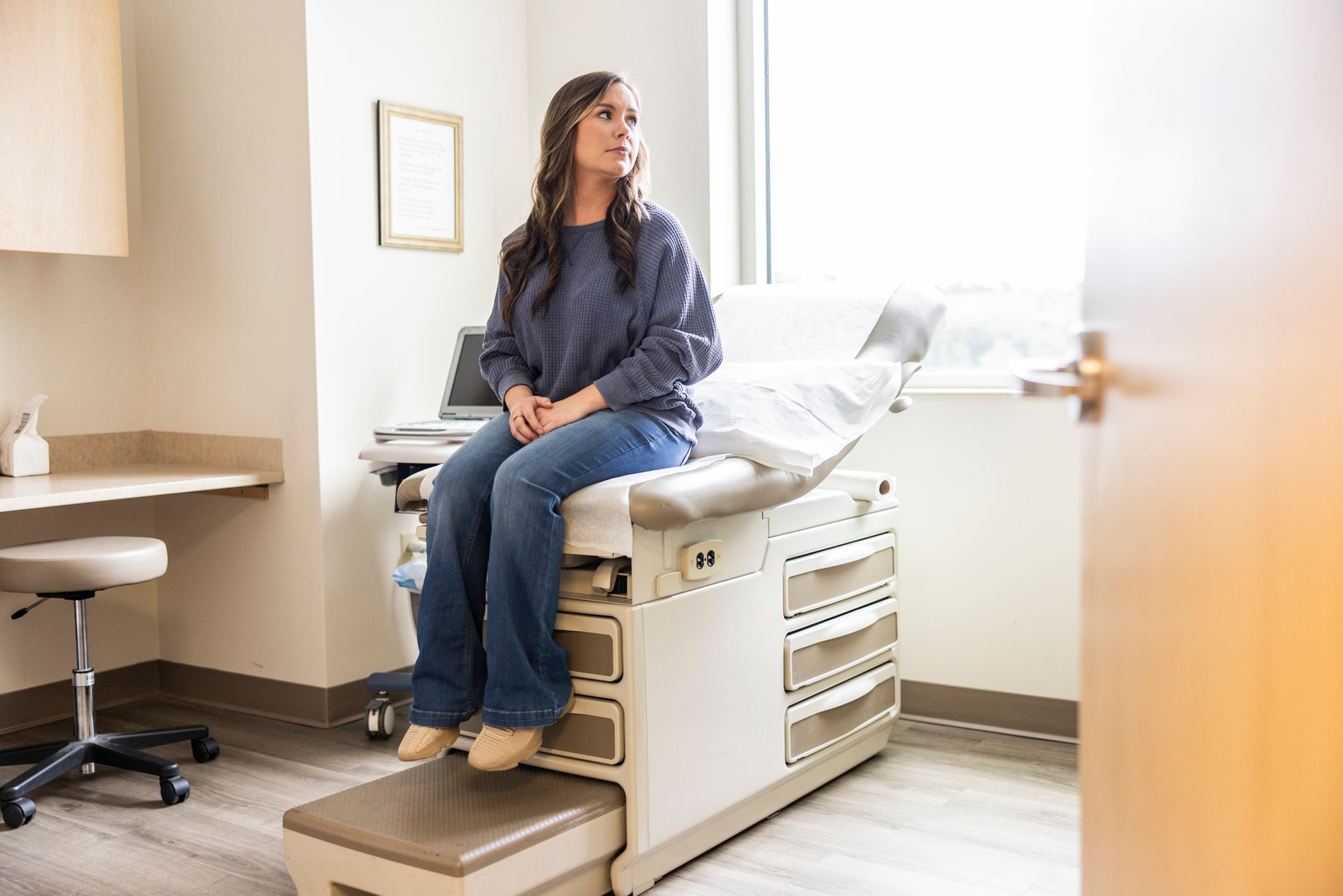 Woman sitting on an examination table in a medical office, looking out the window.