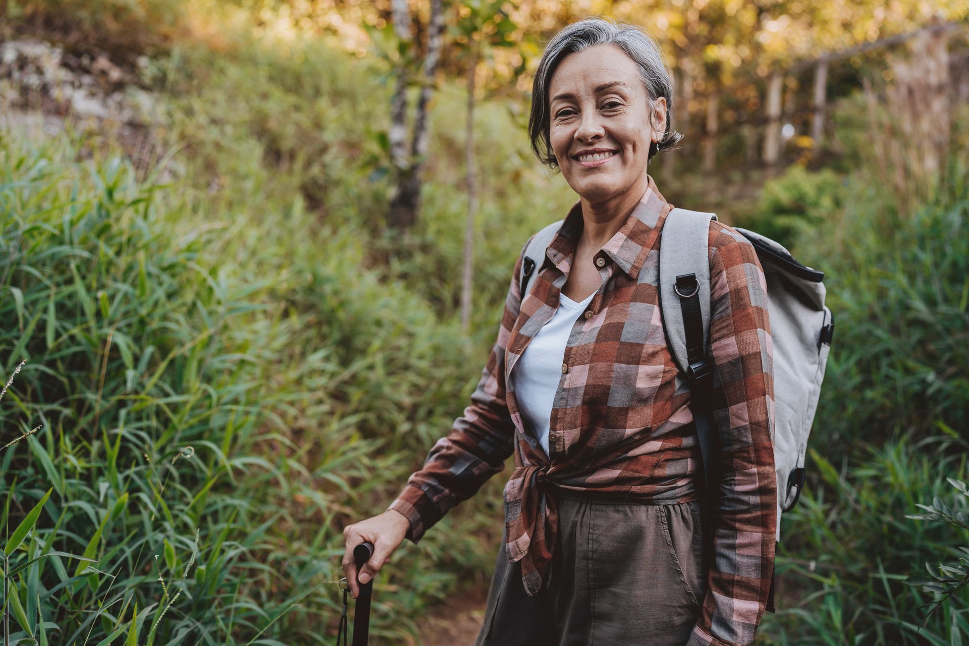 Smiling woman with gray hair hikes in a green field, wearing a backpack and holding a walking stick.