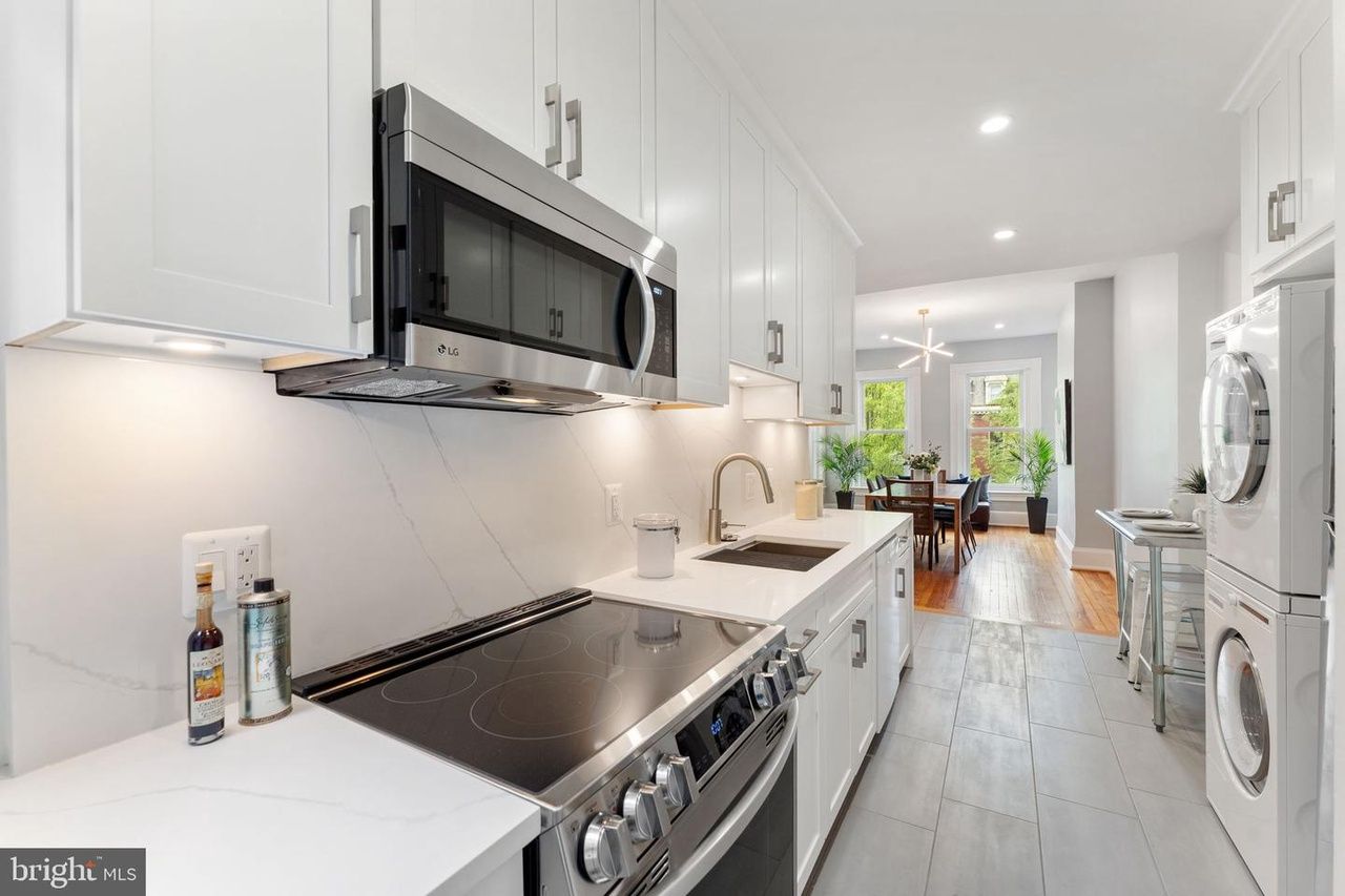 Narrow galley kitchen with white shaker cabinets, quartz backsplash, stainless steel appliances, and stacked washer and dryer near dining area