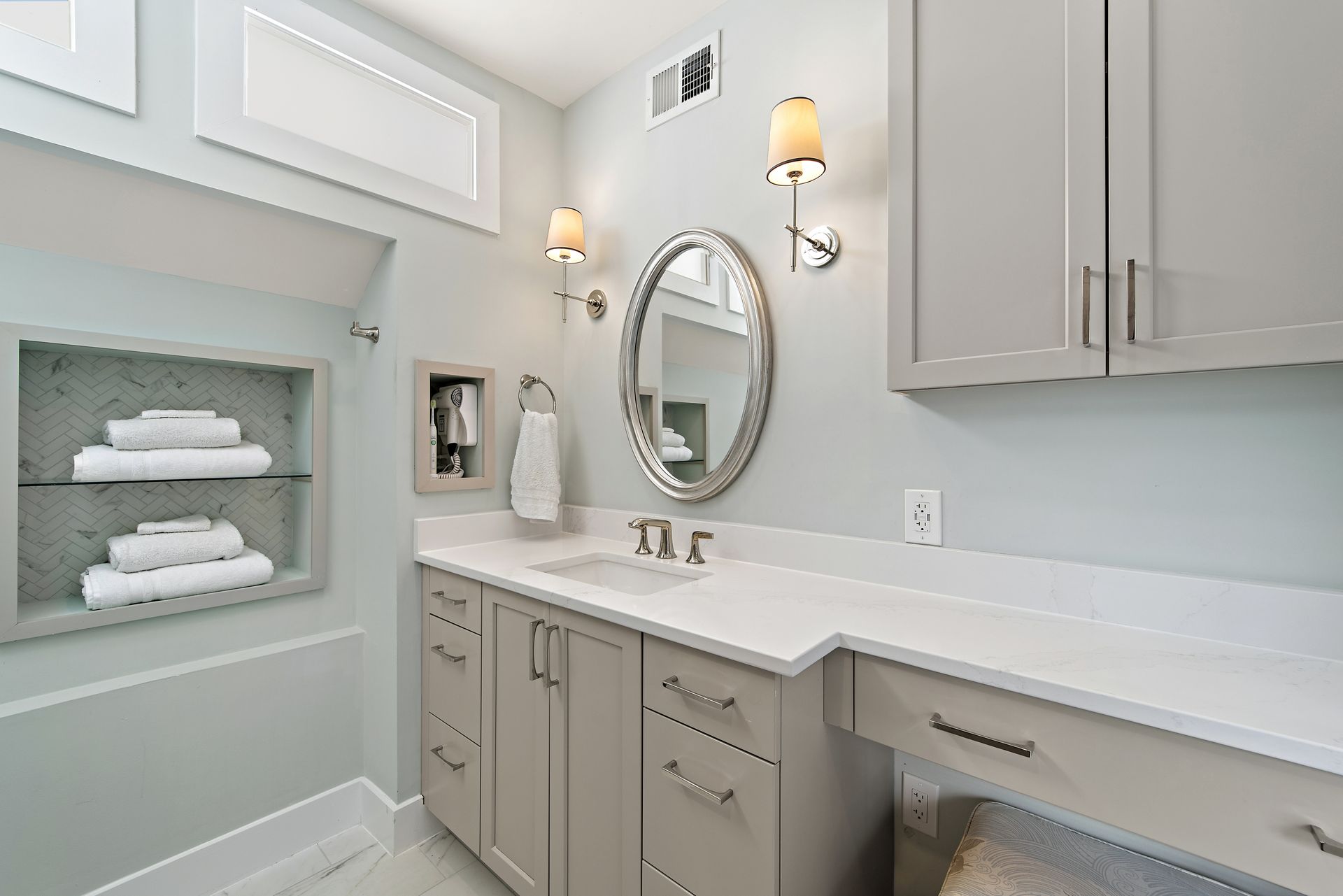 Vanity with makeup counter, round mirror, built-in towel niche, soft gray cabinetry, and herringbone tile accent