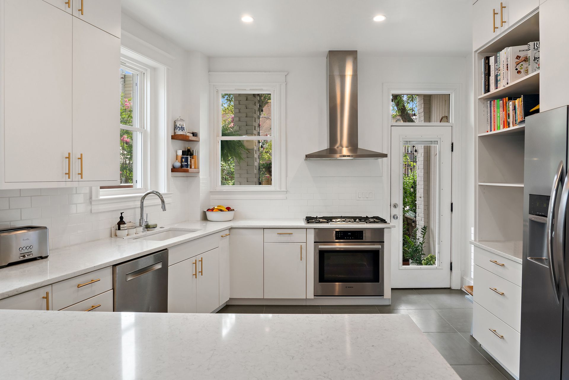 Bright modern kitchen with white cabinets, stainless steel appliances, open shelving, and a glass door leading to a lush backyard.