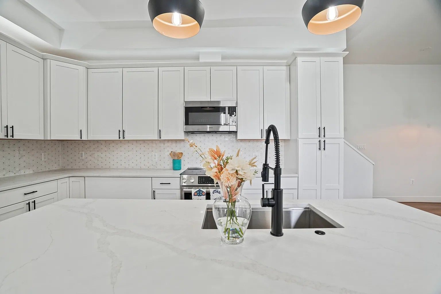 All-white kitchen with shaker cabinets, black hardware, quartz countertops, black pull-down faucet, and floral arrangement on the island