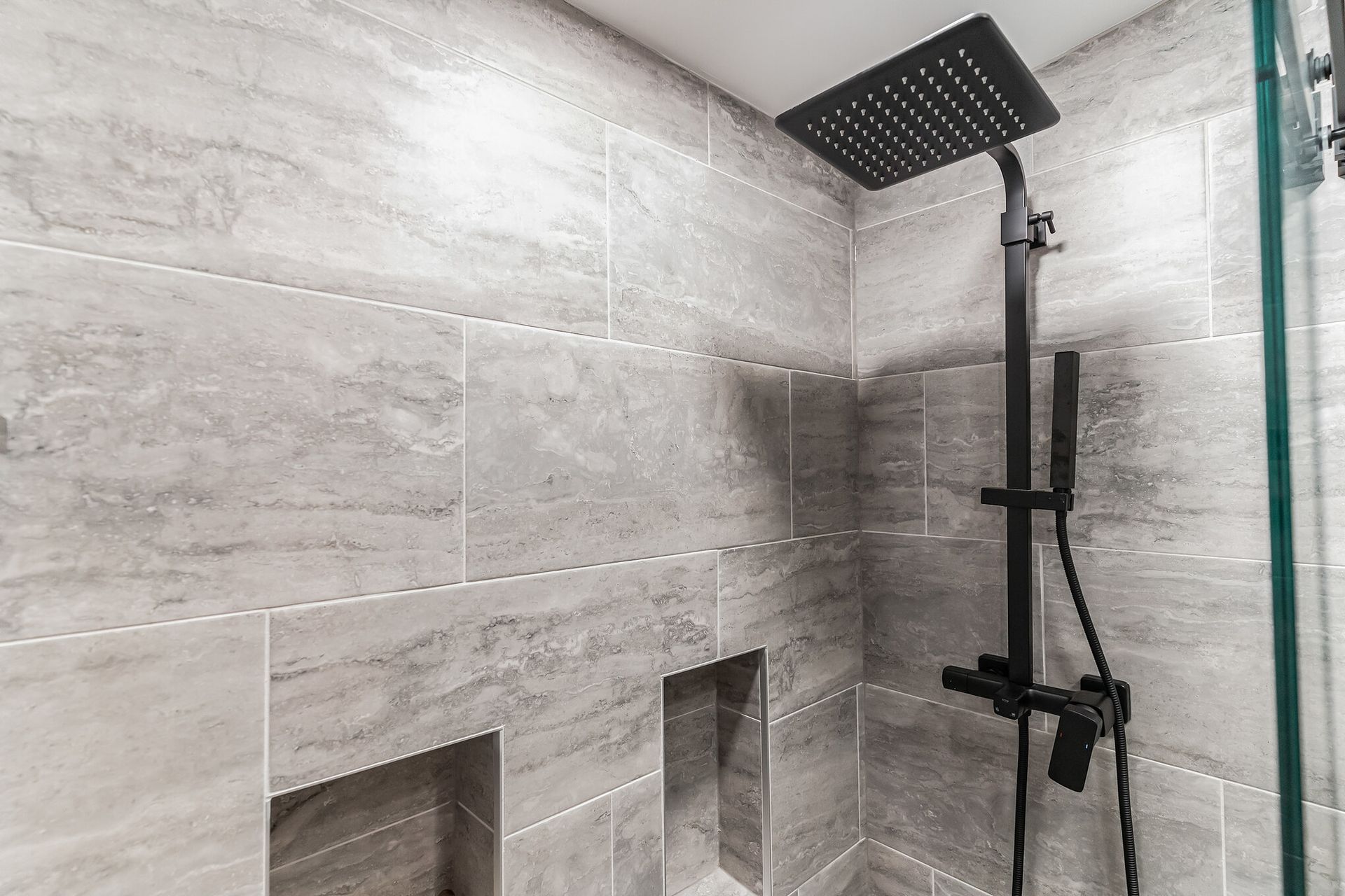 black rainfall shower head with handheld spray and gray stone-look tile walls