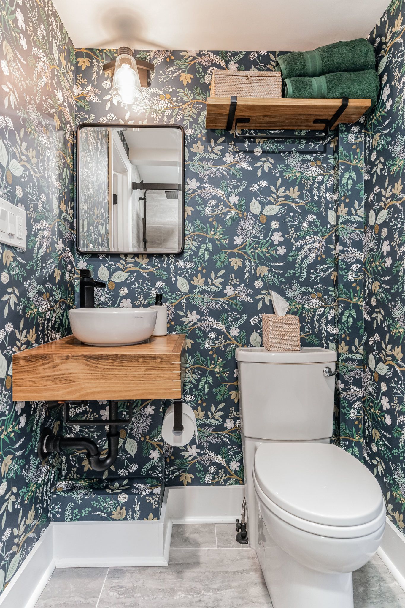 small bathroom with floral wallpaper, wood vanity, vessel sink, and open shelving with green towels