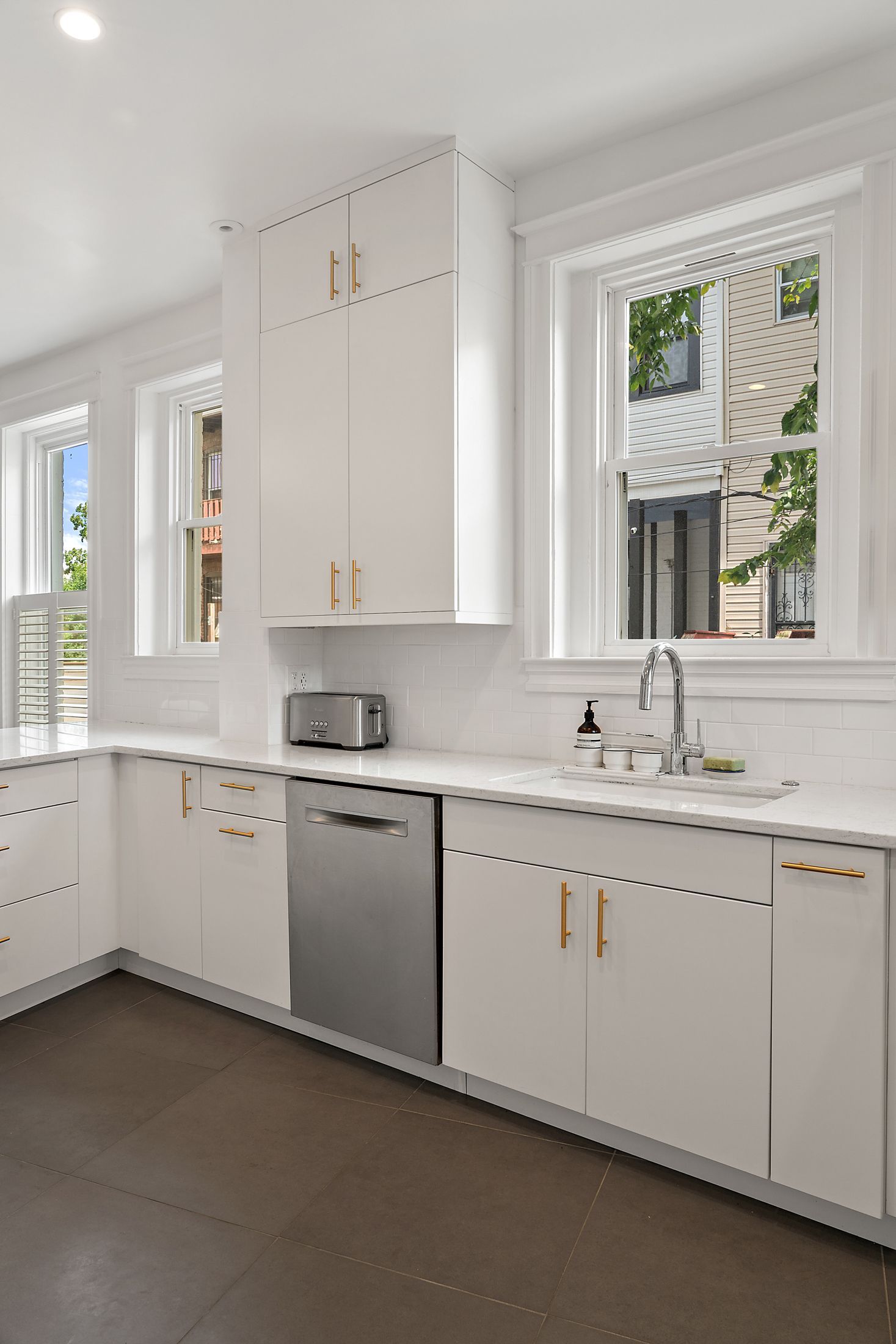 Bright white kitchen with modern gold hardware, stainless dishwasher, quartz countertops, and large double-hung windows above the sink.