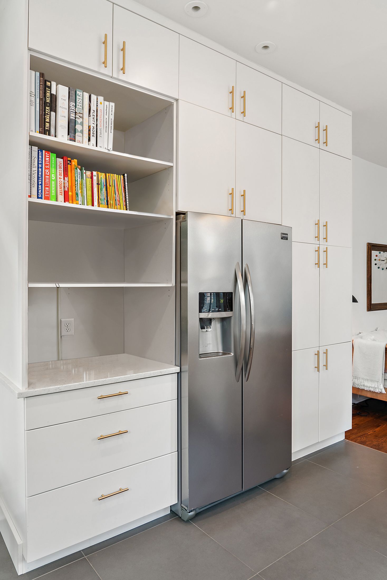 Built-in kitchen storage wall featuring a stainless steel refrigerator, full-height flat panel cabinetry, open bookshelf with cookbooks, and brass hardware