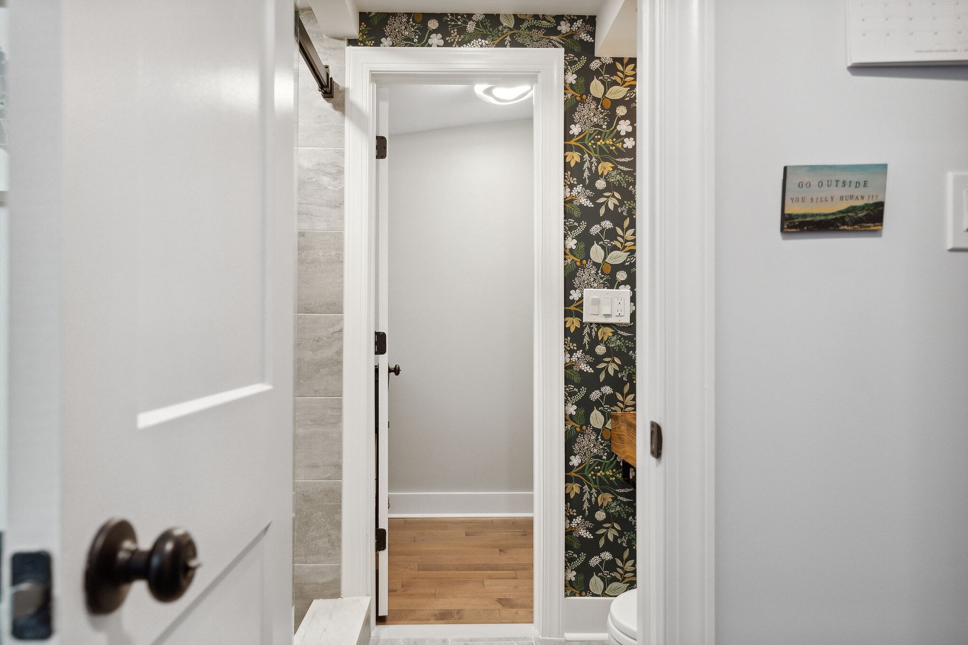 view into floral wallpaper bathroom from hallway with white trim and wood flooring